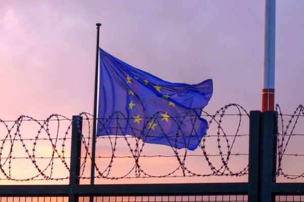 A EU flag behind barbed wire fencing blowing against a sunset sky